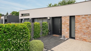 Ein modernes Gebäude mit flachem Dach, einer Steinfassade und einem kleinen Garten mit Hecke und einer kugelförmigen Pflanze.