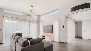 Ein modernes Wohnzimmer mit einem grauen Sofa, einem HolztV-Schrank und großen Fenstern mit transparenten Vorhängen.