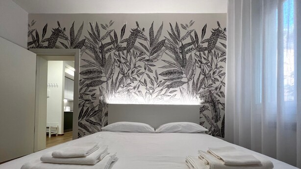 Une chambre à coucher avec un grand lit, du linge de lit blanc, une tête de lit ornée d’un motif de feuilles et une vue sur la salle de bain.