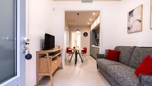 Ein modernes Wohnzimmer mit Sofa, Fernseher auf einem Holzboden, einem Esstisch mit Stühlen und einer Küche im Hintergrund.