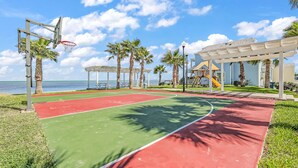 Ein Basketballplatz im Freien mit roter und grüner Oberfläche, umgeben von Palmen und Blick auf den Ozean.