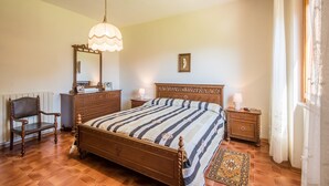Een slaapkamer met een houten bed, een dressoir, een stoel, een nachtkastje, een lamp, een spiegel en een kamerkleed met dessin.
