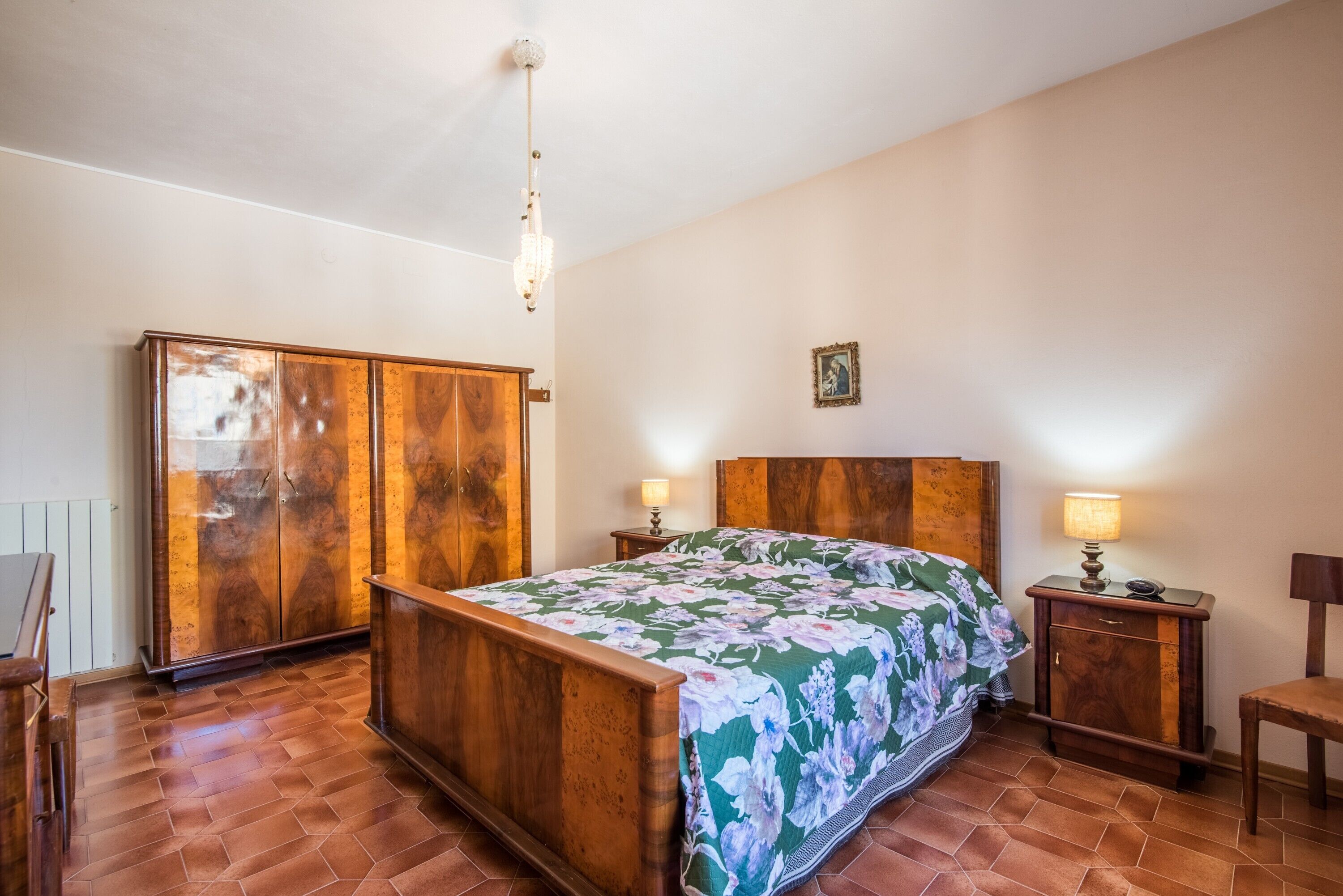 A bedroom with a wooden bed, two bedside tables, a nightstand, a wardrobe, and a chandelier.
