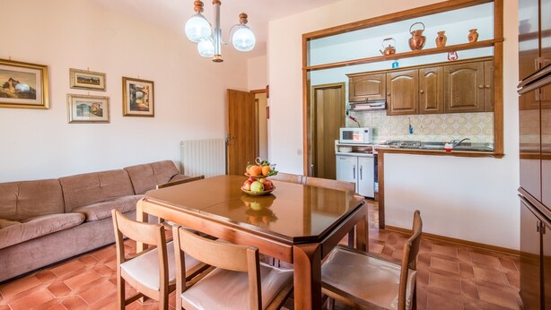 Een eethoek met een houten tafel en stoelen, een bank, en een keuken met houten kastjes en een magnetron.