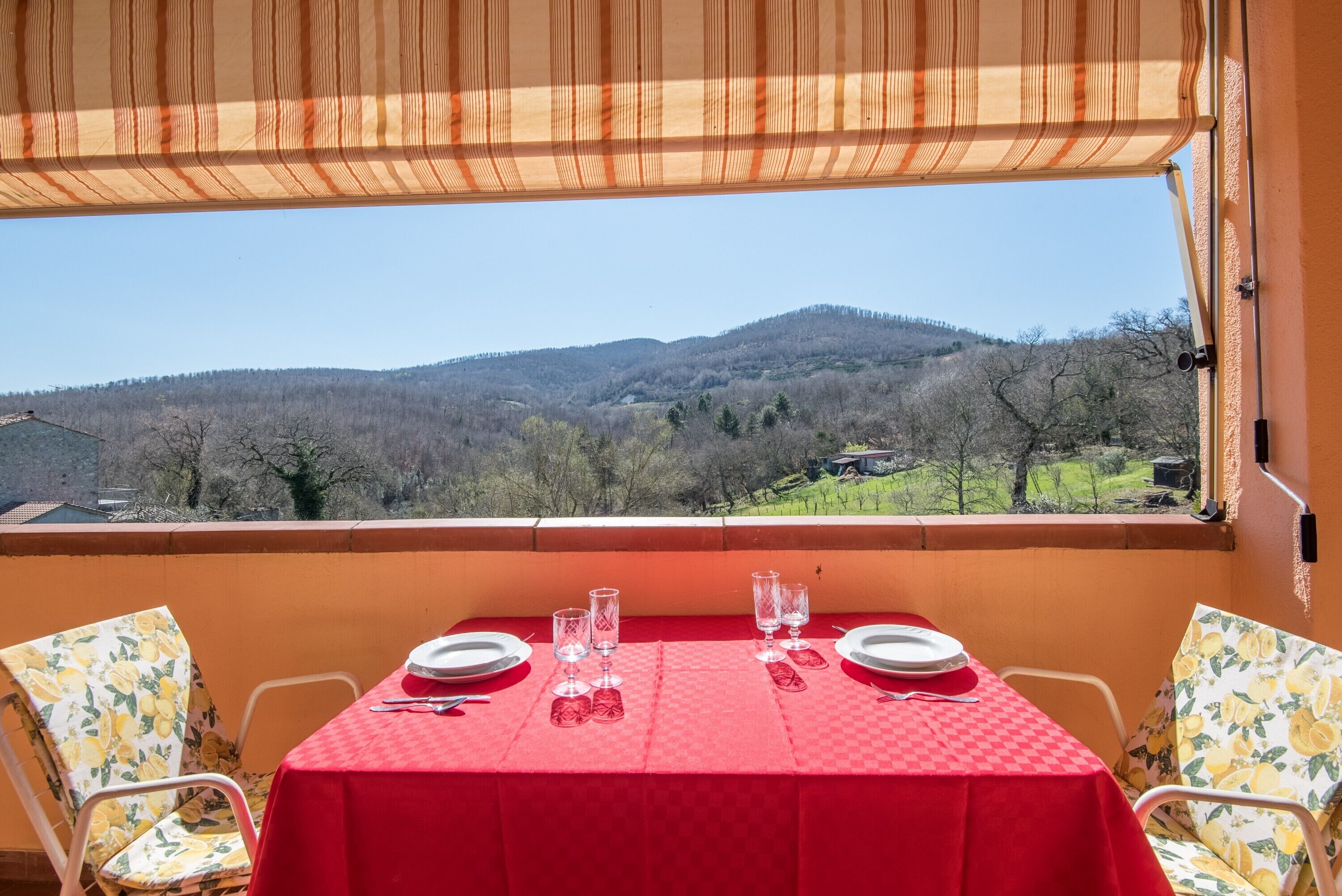 Ein Tisch für zwei, gedeckt mit einer roten Tischdecke, zwei Stühle mit floralem Bezug und Blick auf einen bewaldeten Hügel.