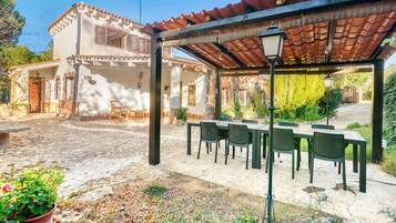 Un espacio exterior cubierto para comer, con una mesa, sillas, una pérgola y un edificio con techo de tejas y ventanas.