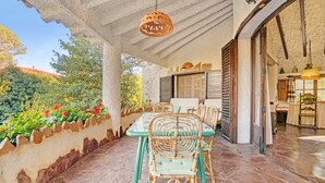 Une terrasse couverte avec une table et des chaises, un luminaire suspendu et une vue sur un jardin où poussent des fleurs rouges.