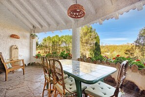 A covered patio with a tiled table and wicker chairs, surrounded by a stone floor and a view of trees and flowers. - Holiday Home 'Rural Kairós' with Mountain View and Wi-Fi (Siete Aguas)