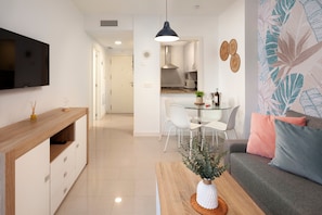 En moderne stue med et fjernsyn, et træskab, en sofa med puder, et sofabord med en plante, samt et spiseområde med stole og et bord.
