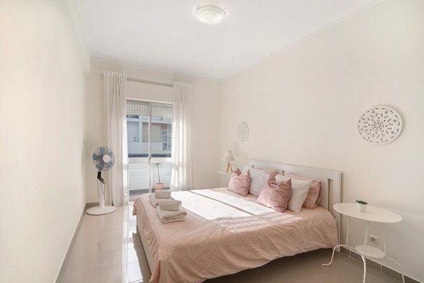 Une chambre à coucher avec un lit, une table de chevet, un ventilateur sur pied et une fenêtre avec des rideaux.