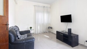 A modern living room with a leather sofa, a flat-screen TV mounted on the wall, a small side table, and a window with sheer curtains. - Apartment 'Matinhos House' with Balcony, Wi-Fi and Air Conditioning (Faro)