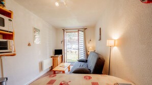 A small, cozy room with a bed, a sofa, a TV, a lamp, and a window with a view of the outside. - Studio "Cabine Ax Les 3 Domaines" in a Quiet Setting Near the Slopes (Ax-les-Thermes)