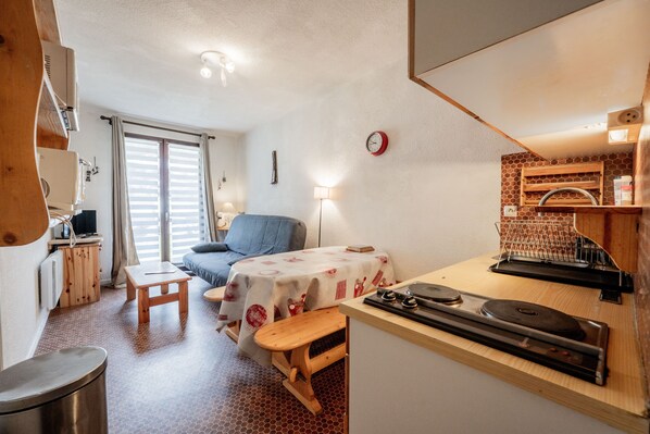 A compact room with a bed, a small table, a bench, a kitchenette, and a window with blinds.