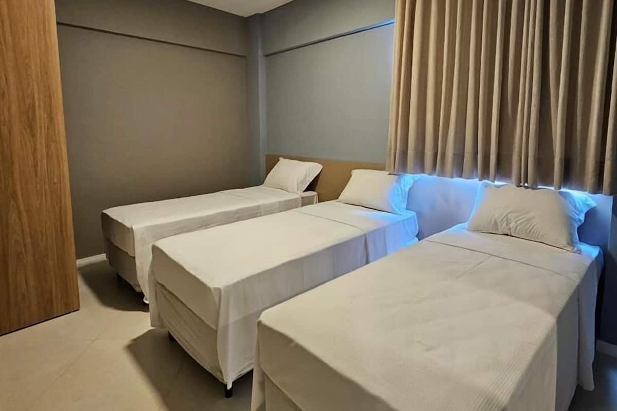 A hotel room with three single beds, a wooden wardrobe, and a large window with curtains.