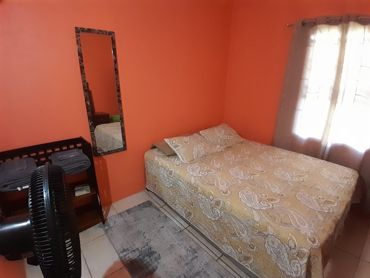 Ein kleiner Raum mit einer orangefarbenen Wand, einem Bett mit gemustertem Überwurf, einem schwarzen Ventilator, einem kleinen Tisch und einem Spiegel.