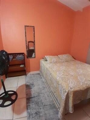 A bedroom with an orange wall, a bed with patterned bedding, a standing fan, and a small wooden bench with folded clothes. - Upstairs 2 bedroom (Castries)