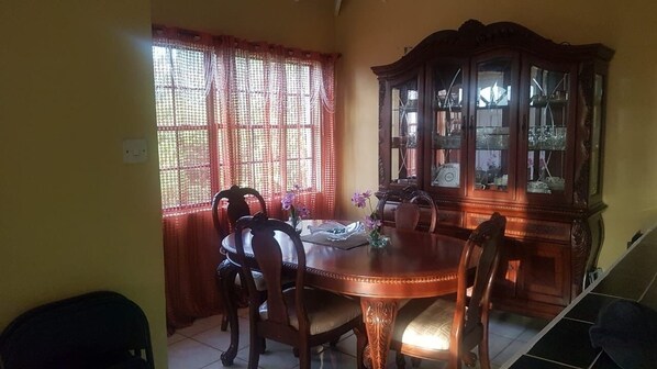 A dining area with a wooden table and chairs, a cabinet with glass doors, and a window with red curtains. - Upstairs 2 bedroom (Castries)