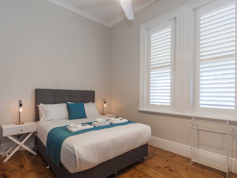 Un dormitorio con cama, mesitas de noche, ventana con persianas y radiador.