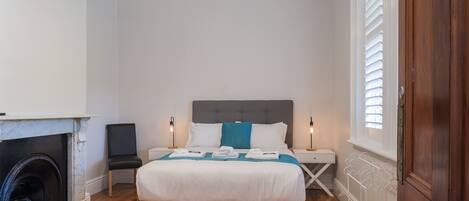 A bedroom with a bed, bedside tables, a fireplace, and a window with blinds.