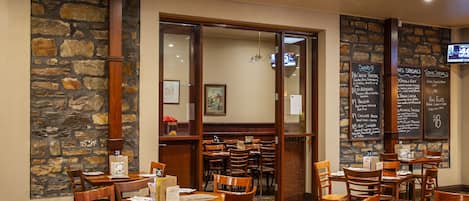 A restaurant with stone walls, wooden tables, and chairs. There are menus on the walls and a TV screen.