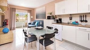Et moderne køkken med hvide skabe, et glasbord i spiseområdet og en blå sofa i opholdsstuen.