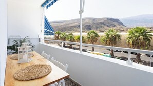 En balkon med et træbord, hvide stole og udsigt til palmer og bjerge.