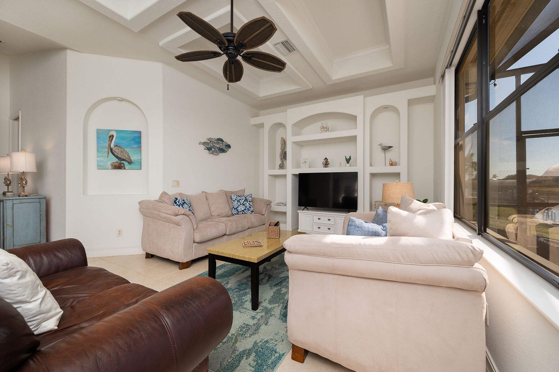 A spacious living room with a beige sofa, a brown leather couch, and a white armchair. There is a ceiling fan, a television, and a large window offering a view of the outdoors.