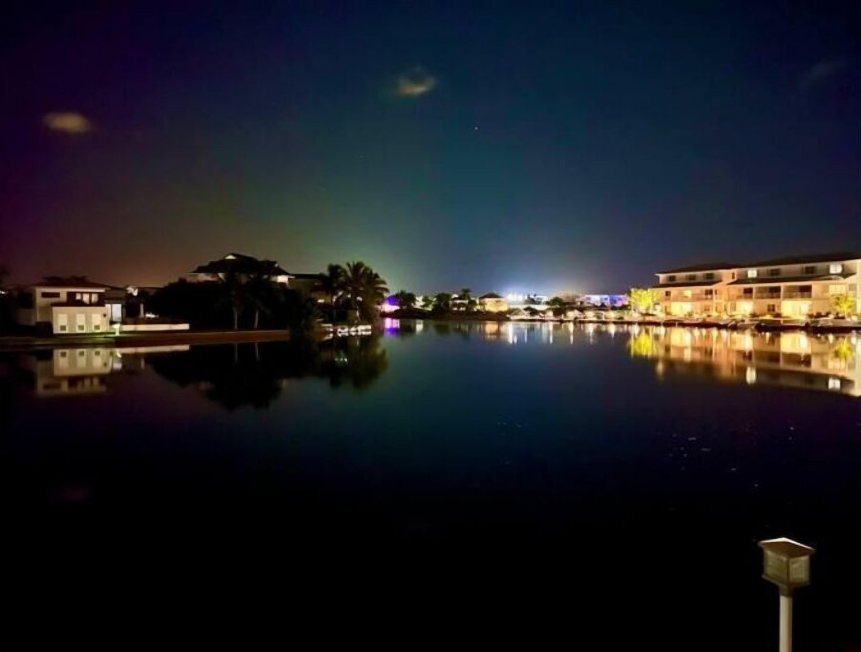 A nighttime view of a waterfront hotel with a calm pool reflecting the buildings and a visible aurora.