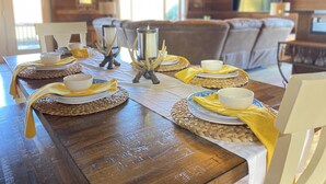 Una mesa de comedor de madera dispuesta con platos blancos, tazones y servilletas amarillas. Se observan candelabros decorativos y individuales tejidos.