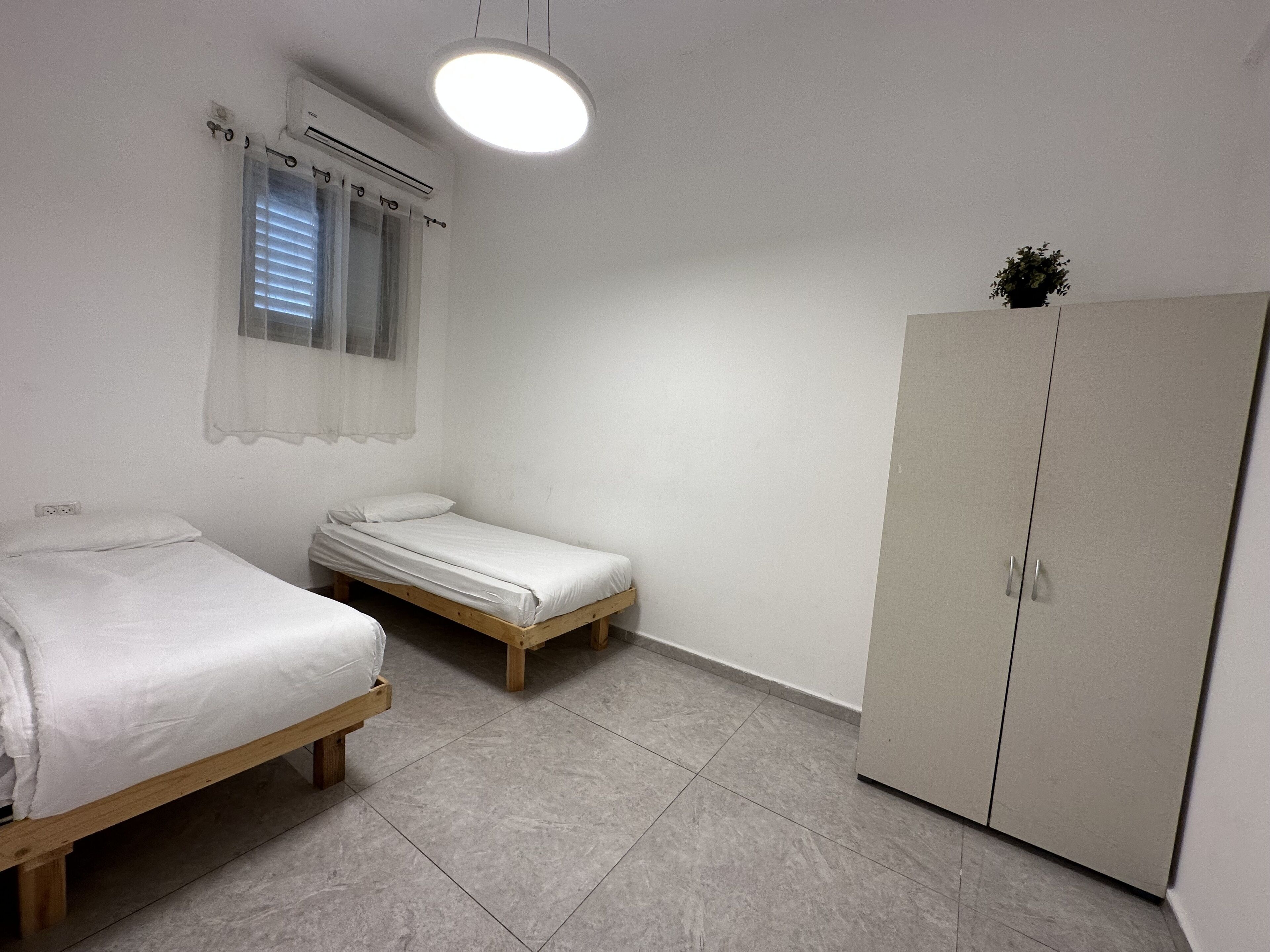A small, single-room accommodation with two single beds, a wardrobe, a window with curtains, and a ceiling light.