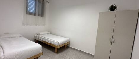 A small, single-room accommodation with two single beds, a wardrobe, a window with curtains, and a ceiling light.