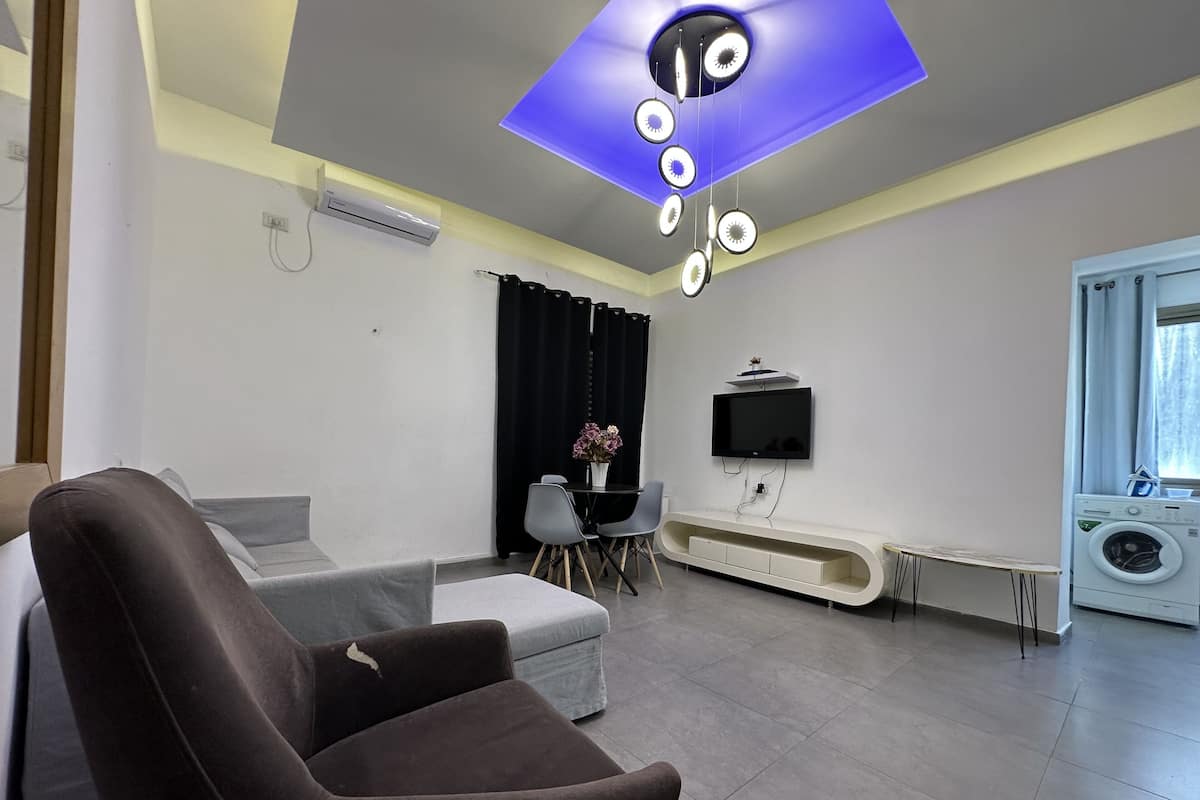 Un salon moderne avec un canapé, un téléviseur fixé au mur, un lave-linge et un luminaire suspendu au plafond.