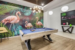 Una mesa de billar en una habitación con un mural tropical, una lámpara moderna y un letrero colocado en la pared.