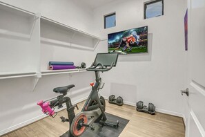 A room with a stationary bike, a wall-mounted TV, and shelves with exercise equipment. - Flamingo Palms: Pool, Spa, Pet-Friendly, Games! (Indio)