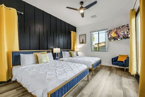 A hotel room with two beds, a ceiling fan, a window with a city view, and a blue armchair. - Flamingo Palms: Pool, Spa, Pet-Friendly, Games! (Indio)