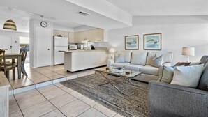 En moderne stue med sofa, sofabord og et tæppe. Der er et køkkenområde med skabe og hårde hvidevarer i baggrunden.