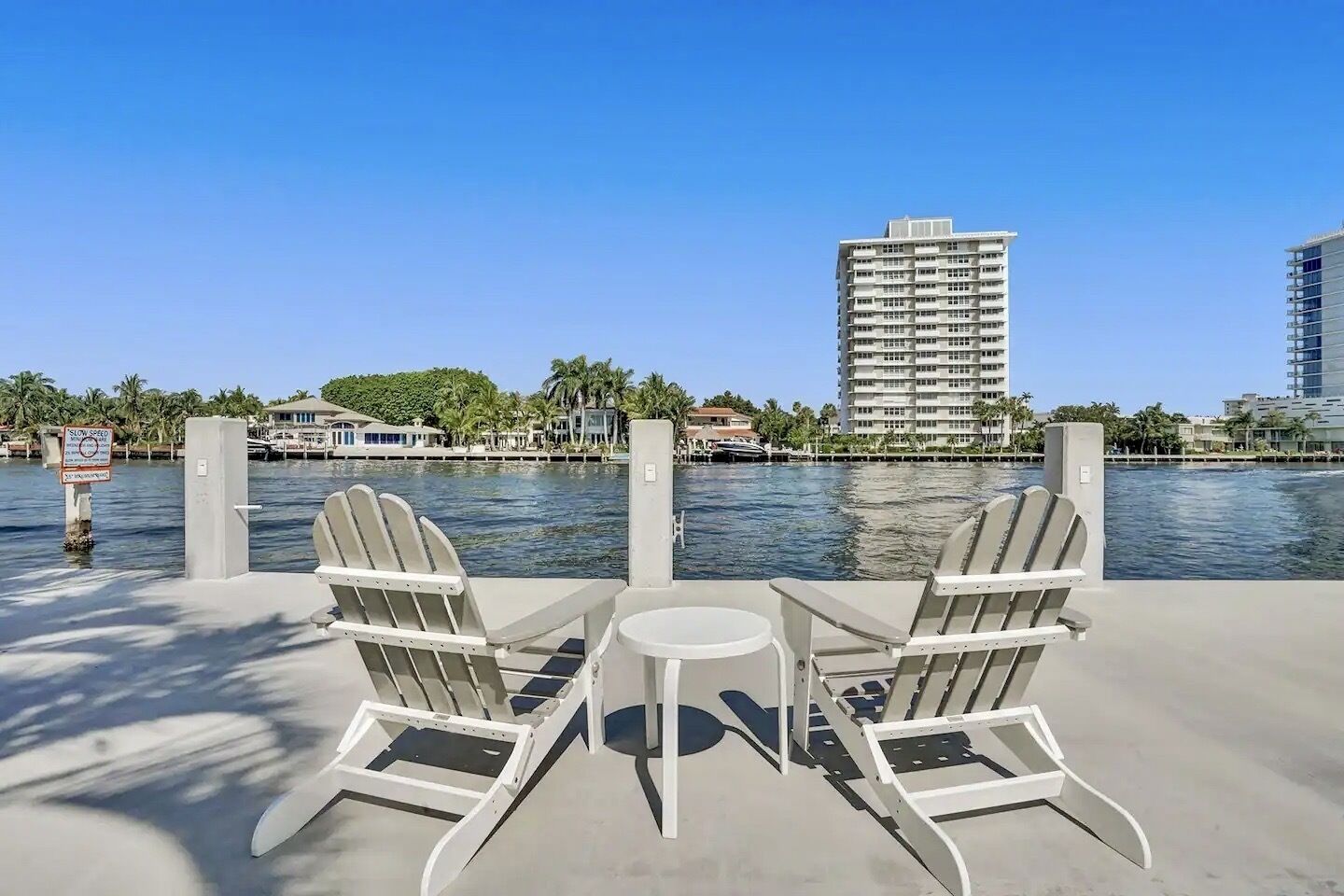 A waterfront patio with two Adirondack chairs and a small table, overlooking a body of water with a 'SLOW SPEED' sign and a tall building in the background.