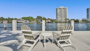 Une terrasse au bord de l’eau, avec deux fauteuils Adirondack et une petite table, donnant sur une étendue d’eau où l’on peut voir un panneau indiquant une « VITESSE RÉDUITE » et, au loin, un grand immeuble.