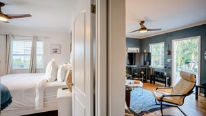 Une chambre à coucher avec un lit, une table de chevet blanche et un ventilateur de plafond. La pièce est dotée d’une grande fenêtre avec des stores et offre une vue sur la verdure à l’extérieur.