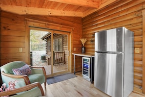 L’intérieur d’un chalet en bois, avec une porte-fenêtre coulissante donnant sur l’extérieur, une petite cuisine équipée d’un réfrigérateur, une chaise avec un coussin à motifs et une terrasse en bois visible à travers la porte.