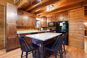 L’intérieur d’un chalet en bois comprend une cuisine équipée de meubles en bois, d’un réfrigérateur noir, d’un four à micro-ondes et d’un îlot en marbre agrémenté de deux tabourets de bar.