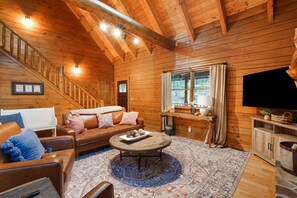 A cozy cabin interior with a wooden staircase, a leather sofa, a coffee table, and a television.