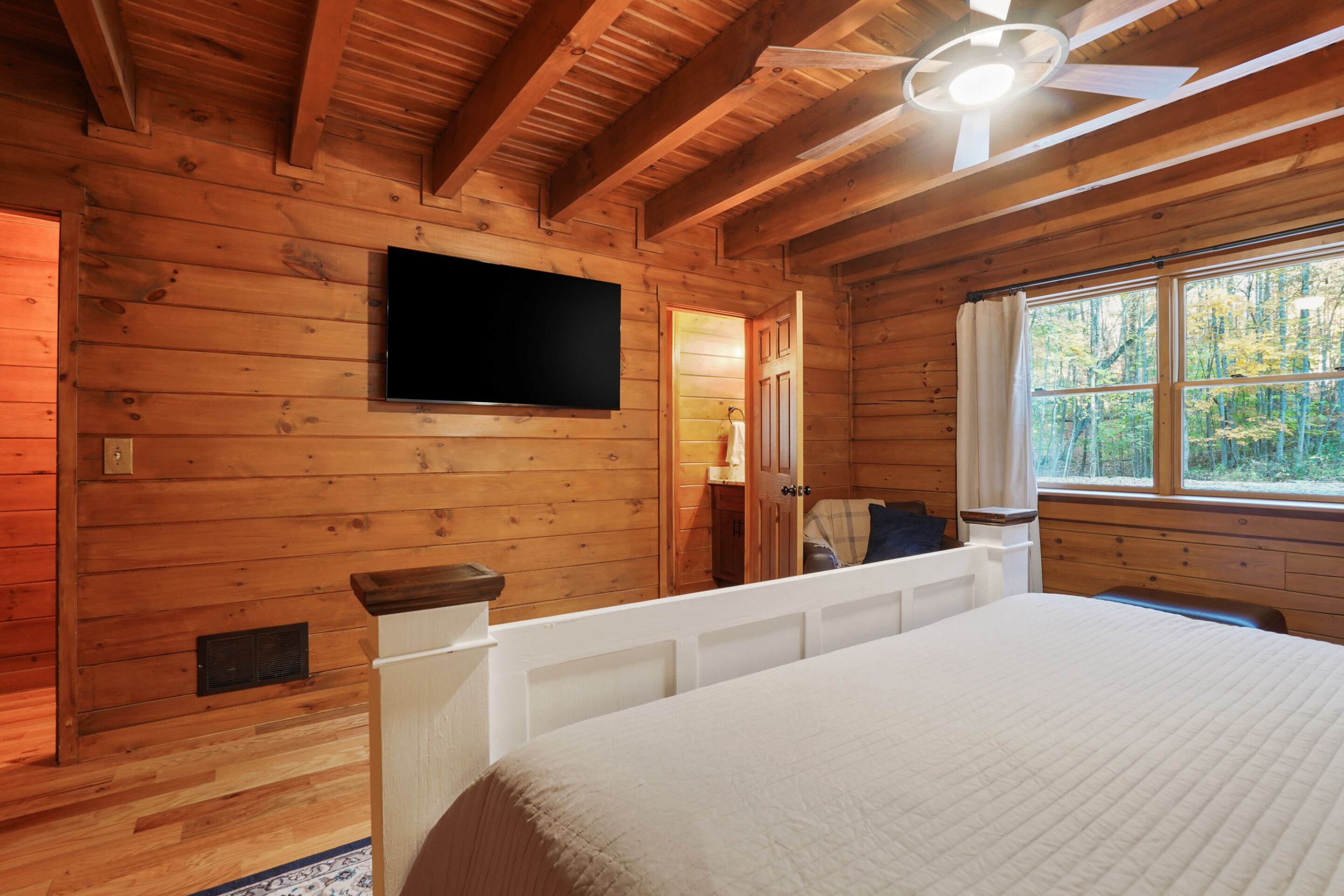 A wooden cabin room with a bed, a mounted TV, a window with a view of trees, and a ceiling fan.