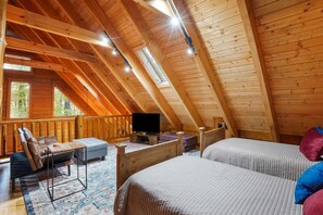 A cozy attic bedroom with wooden beams, a television, two beds, a desk, and a cushioned chair.
