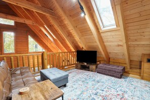 A cozy attic room with a wooden ceiling, a large window, a flat-screen TV, a sofa, an ottoman, and a patterned rug.