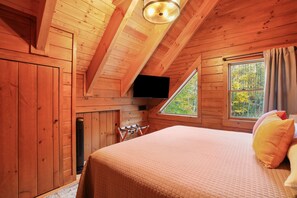 A cozy wooden cabin bedroom with a bed, a TV mounted on the wall, and a window with curtains.