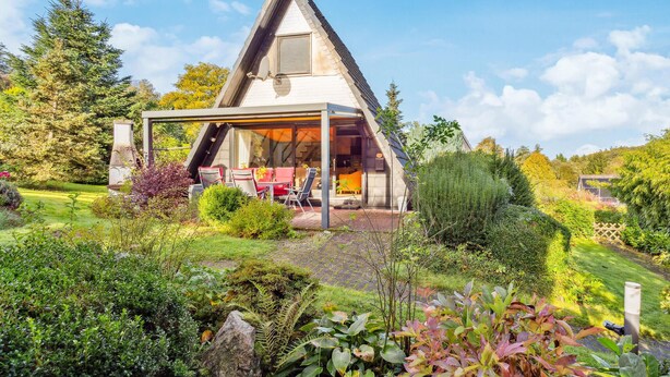 A modern, triangular-roofed house with a covered patio area, surrounded by lush gardens and trees. - Cozy home in Bad Wünnenberg with WiFi (Bad Wünnenberg)