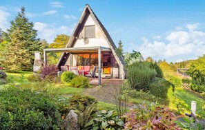 Ein modernes Haus mit dreieckigem Dach und einer überdachten Terrasse, umgeben von üppigen Gärten und Bäumen.