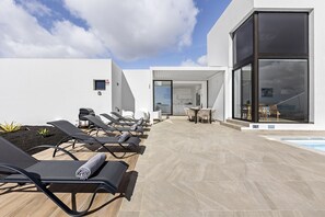 A modern outdoor pool area with sun loungers, a dining area, and a clear view of the sky.