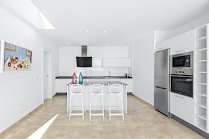 A modern kitchen with white cabinetry, a stainless steel refrigerator, and a built-in oven. There are three white bar stools at the kitchen island.